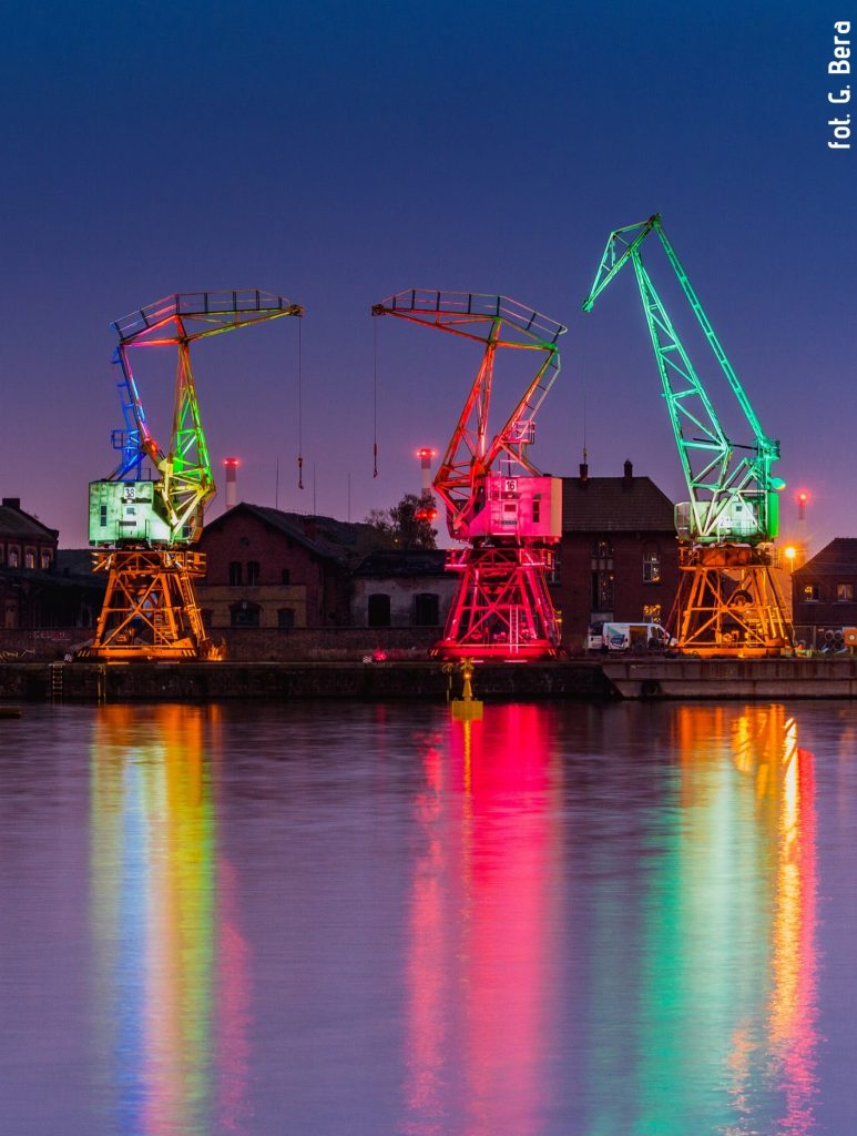 Zdjęcie wykonane przez Grzegorza Berę. Przedstawia trzy podświetlone na kolorowo dźwigi-żurawie stoczniowe na szczecińskiej Łasztowni, przed nimi widok na rzekę Odrę.