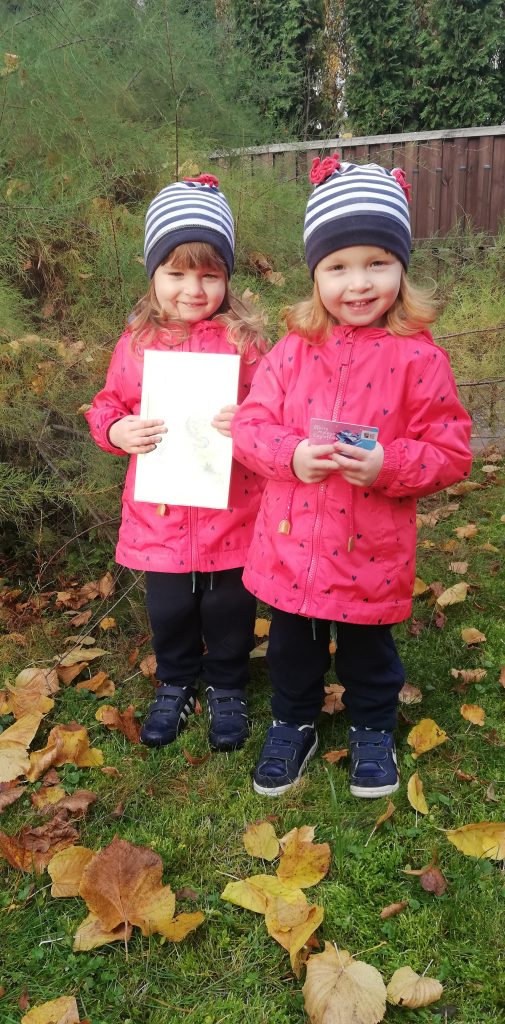 Dwie uśmiechnięte małe dziewczynki. Jedna z nich trzyma dyplom małego czytelnika, druga kartę do zbierania naklejek.