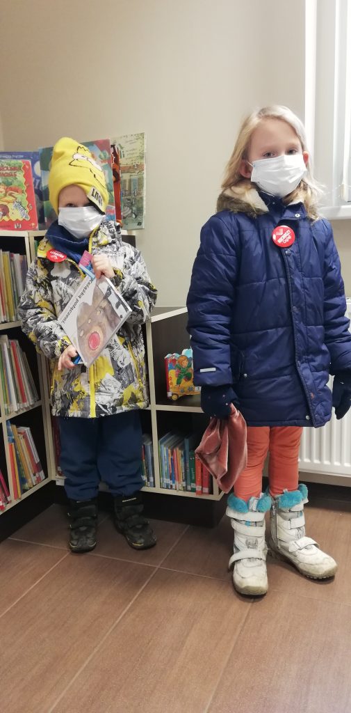 Chłopiec i dziewczynka w maseczkach na twarzy, stoją przed regałem z książkami w bibliotece. Chłopiec trzyma książkę dla dzieci.
