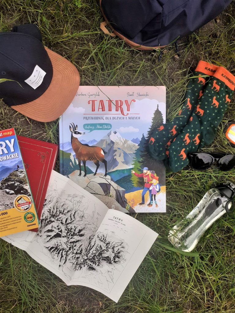 Książka "Tatry: przewodnik dla dużych i małych" leży na trawie. Obok niej butelka z wodą, czapka z daszkiem, skarpety, okulary przeciwsłoneczne i przewodniki po Tatrach
