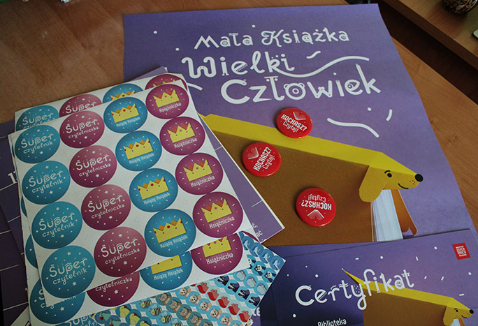 Na stole leżą kolorowe plakaty promujące akcję "Mała książka, wielki człowiek", czerwone przypinki z napisem "Kochasz? Czytaj!" oraz okrągłę, kolorowe naklejki z napisami: Super czytelnik, Książe Książek, Księżniczka.