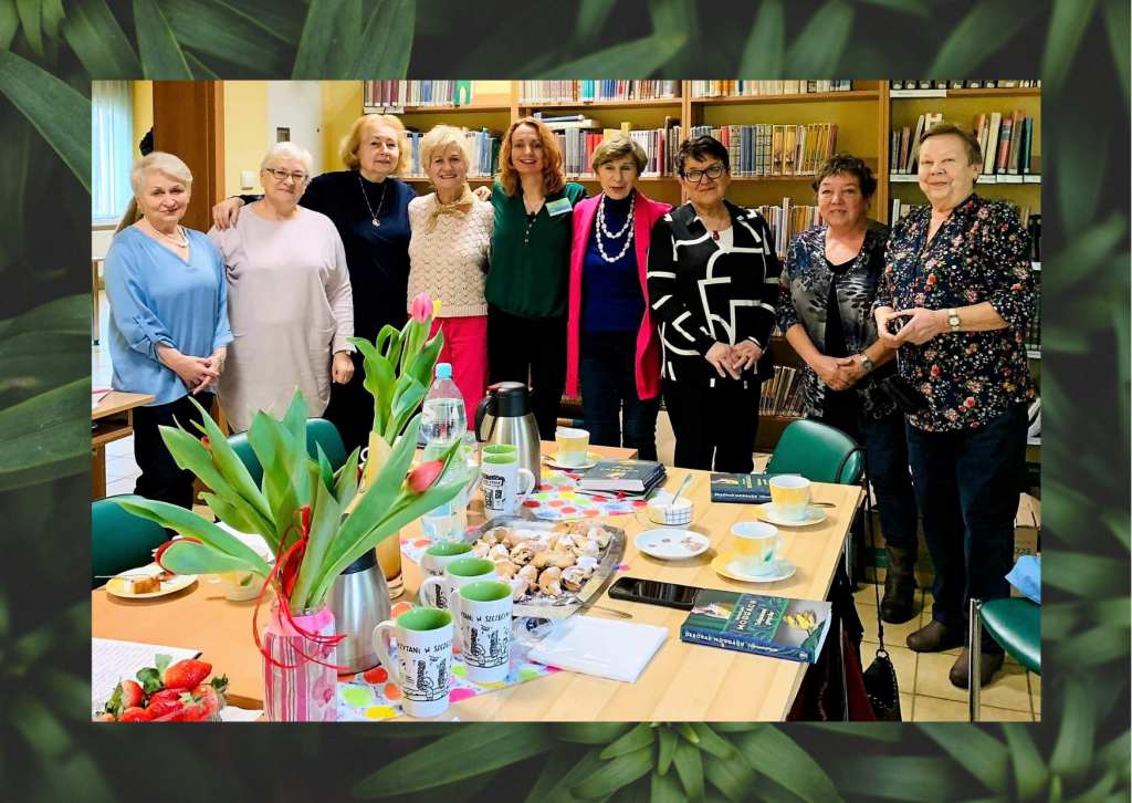 Uśmiechnięte panie z Dyskusyjnego Klubu Książki pozują do zdjęcia w bibliotece. Przed nimi stół, na którym są kwiaty w wazonie i słodycze.
