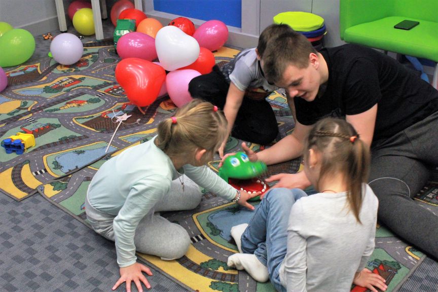 dwie dziewczynki i chłopiec siedzą na dywanie, z nimi nastoletni wolontariusz, bawią się, za nimi koloroqe balony w kształcie serca